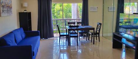 A living room with a blue sofa, a dining table with chairs, a TV, and a balcony with curtains.