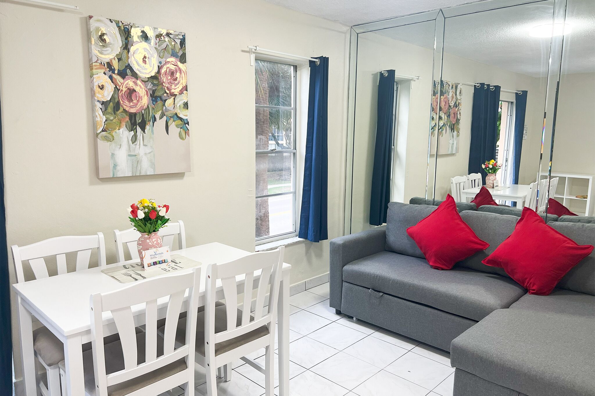 A room with a dining table, white chairs, a grey sofa with red cushions, a large mirror, and a painting of flowers on the wall.