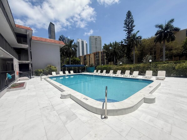 A rectangular pool with a blue border, surrounded by white lounge chairs and a blue canopy.