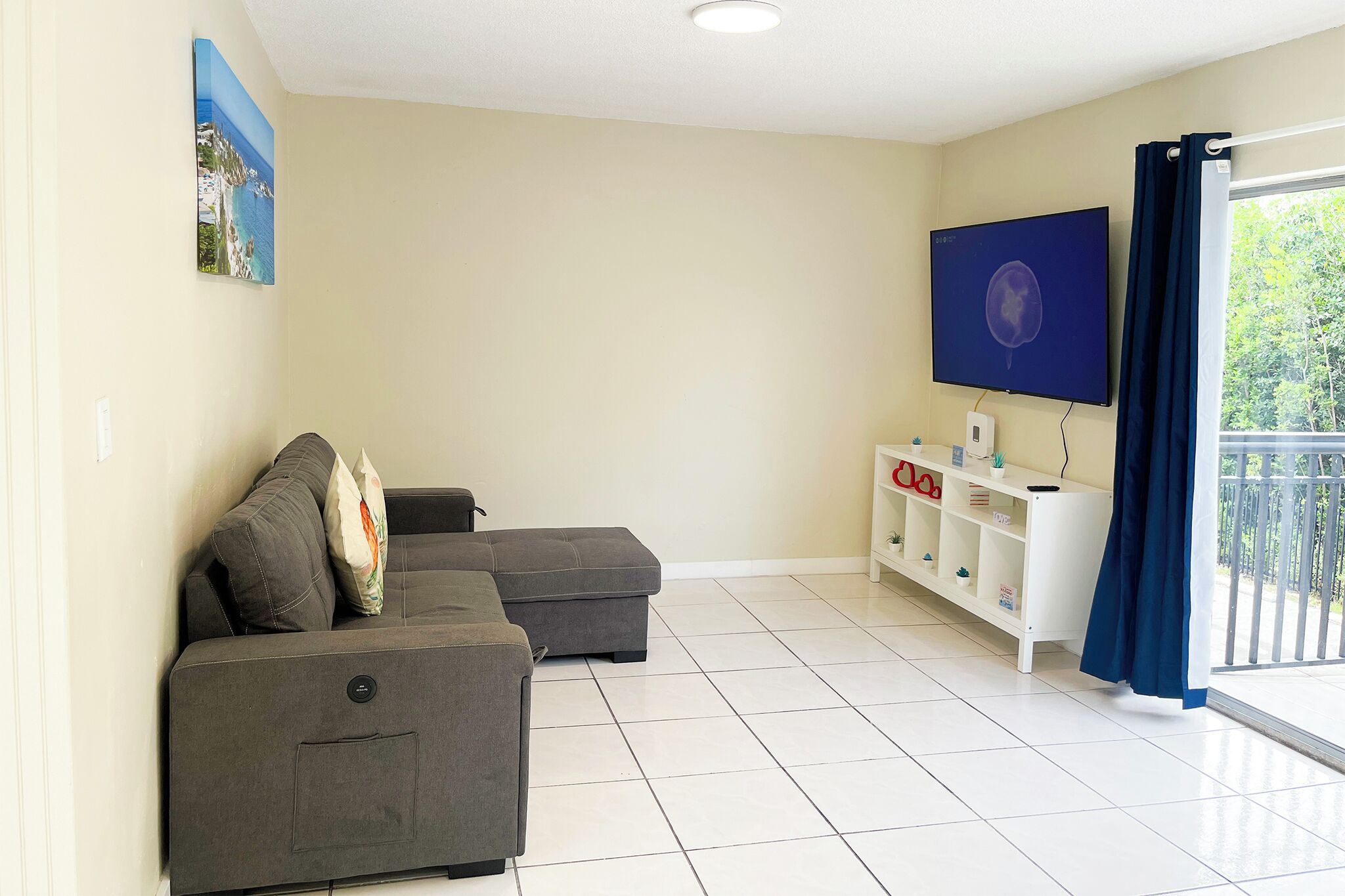 A living room with a grey sectional sofa, a flat-screen TV, a white entertainment unit, and a balcony with a view of greenery.