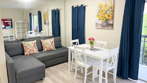 A modern living room with a grey sectional sofa, white chairs, a small dining table, and a large mirror reflecting the room. - Sunny Isles Apartments by MiaRentals (Sunny Isles Beach)