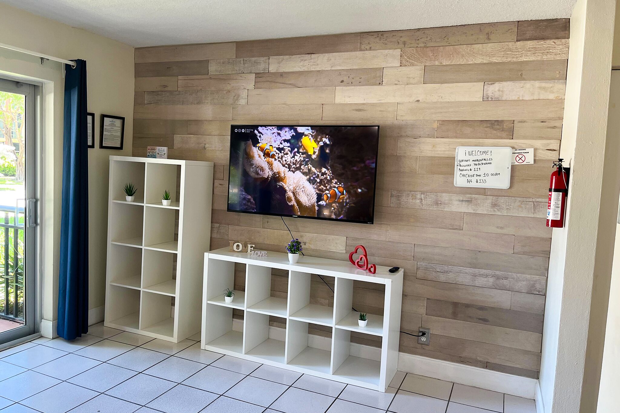 Una sala de estar con un televisor de pantalla plana montado en una pared de madera, un mueble blanco de entretenimiento debajo y un cartel de bienvenida.