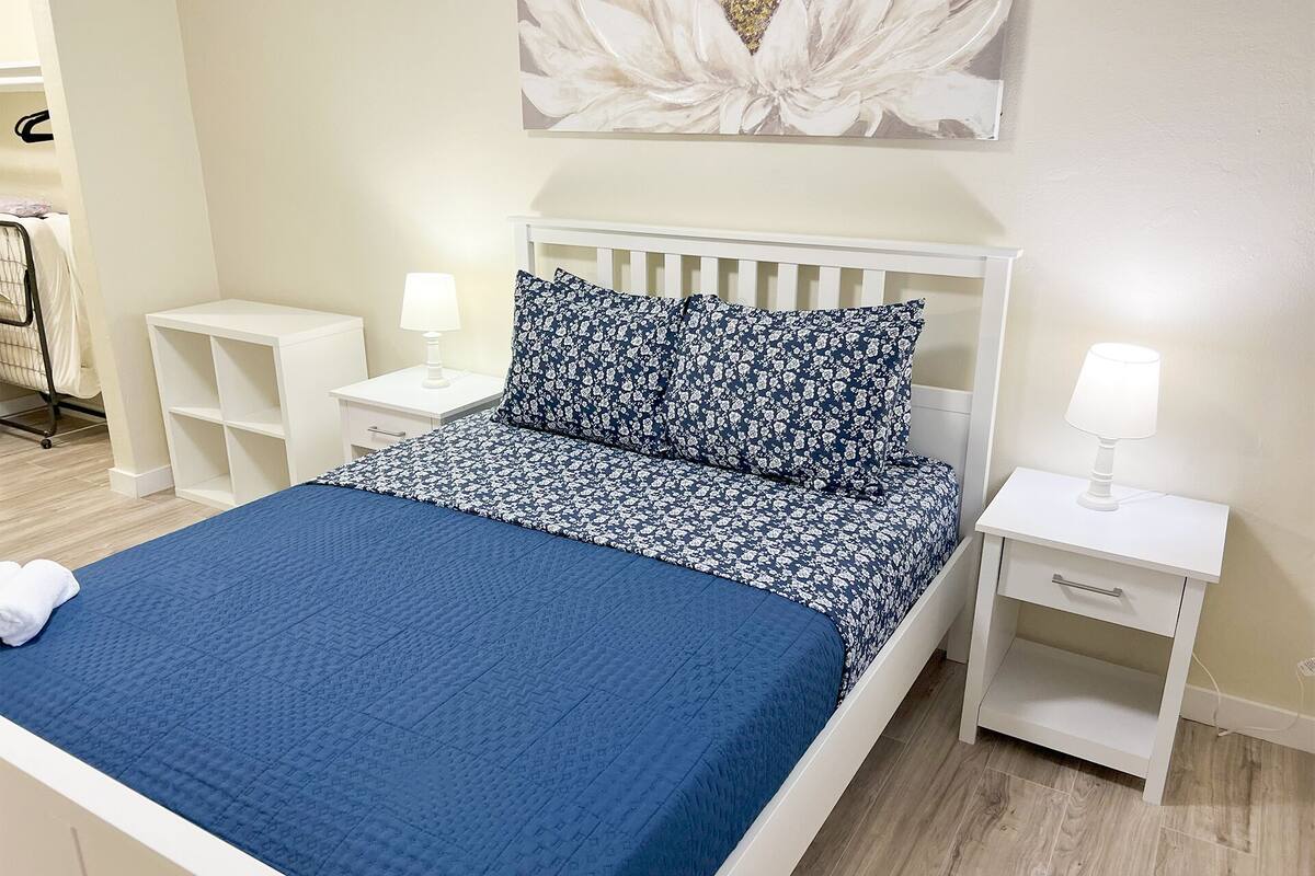 En snyggt bäddad säng med ett blått täcke, två vita sängbord med lampor, en vit sänggavel och ett blommönstrat konstverk som hänger på väggen.