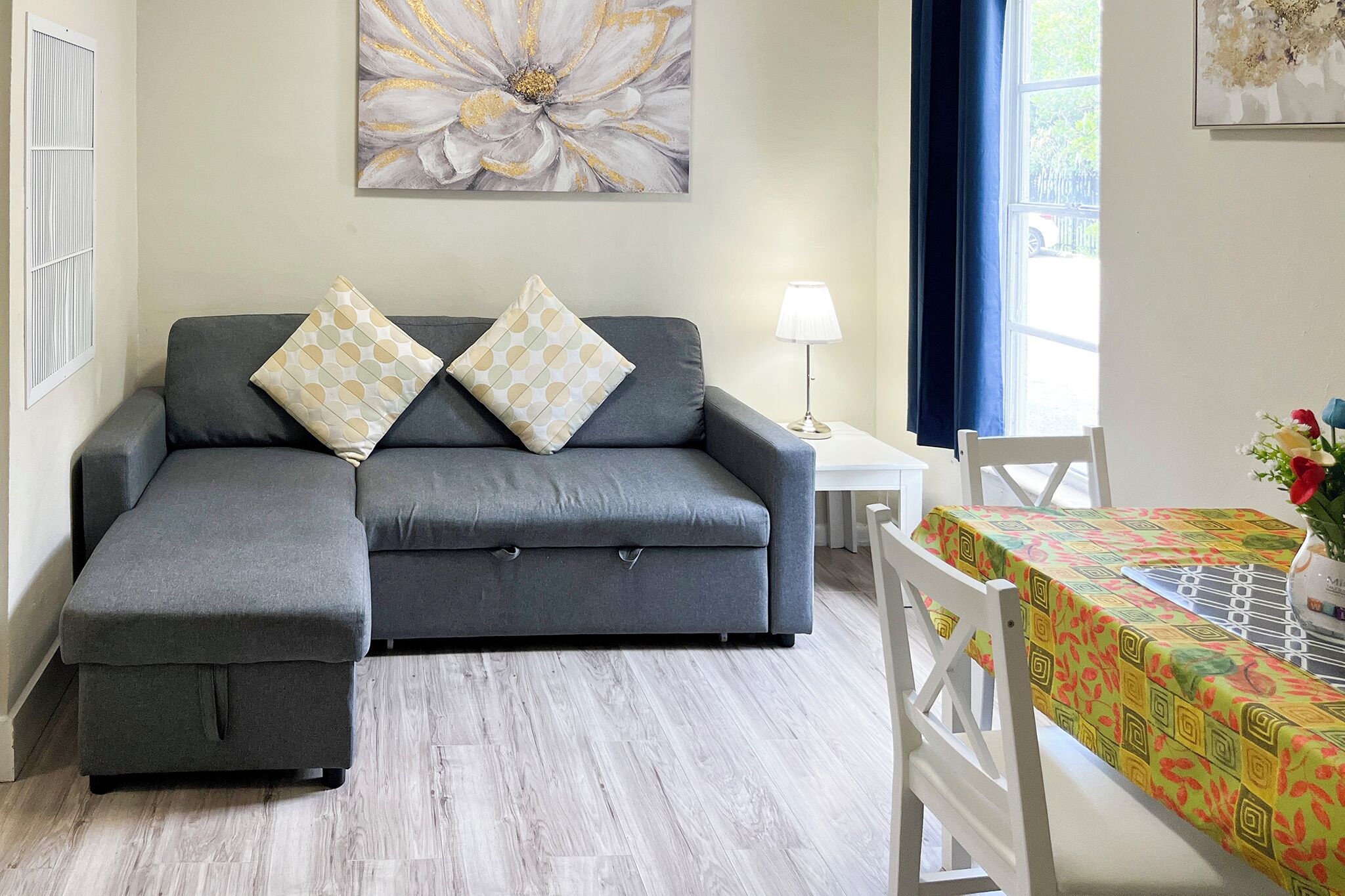 A modern living room with a grey sectional sofa, two patterned cushions, a side table with a lamp, and a dining area with a table covered in a colorful tablecloth.