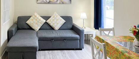 A modern living room with a grey sectional sofa, two patterned cushions, a side table with a lamp, and a dining area with a table covered in a colorful tablecloth.