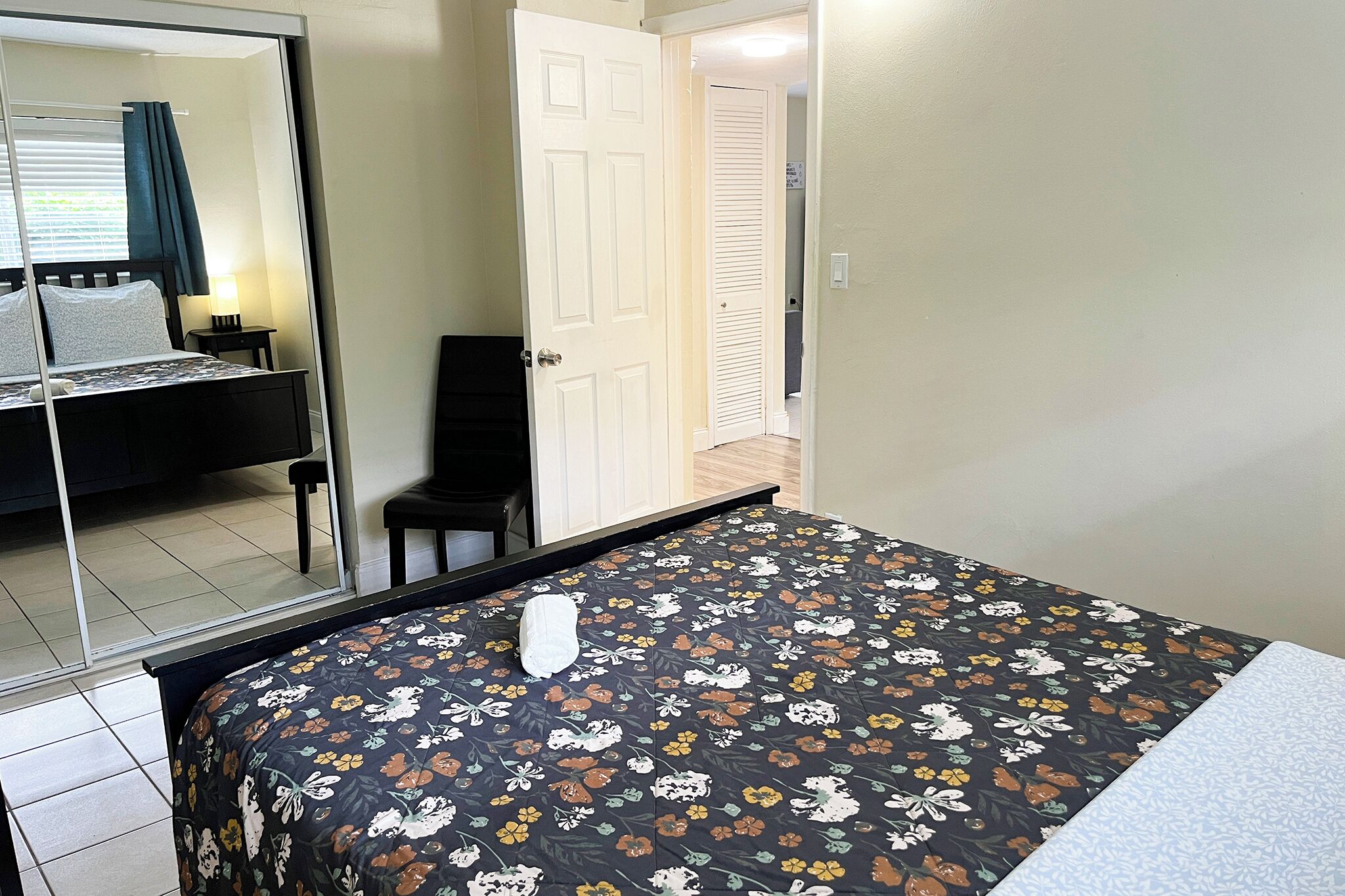A hotel room with a bed, a chair, a mirror, and a door leading to another room.