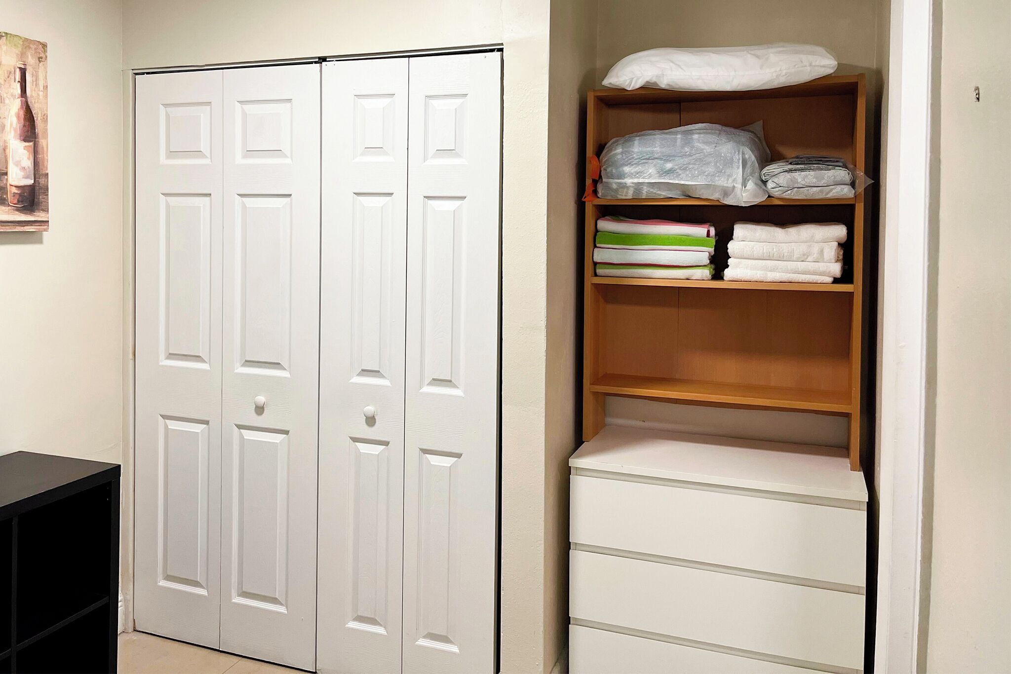 Una habitación con un armario blanco, un estante de madera con toallas dobladas y un mueble de almacenamiento negro.