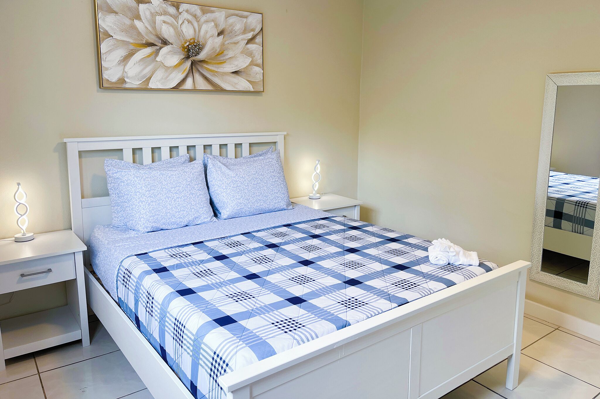 A neatly made bed with a checkered bedspread, two pillows, a bedside table with a lamp, and a wall-mounted mirror.