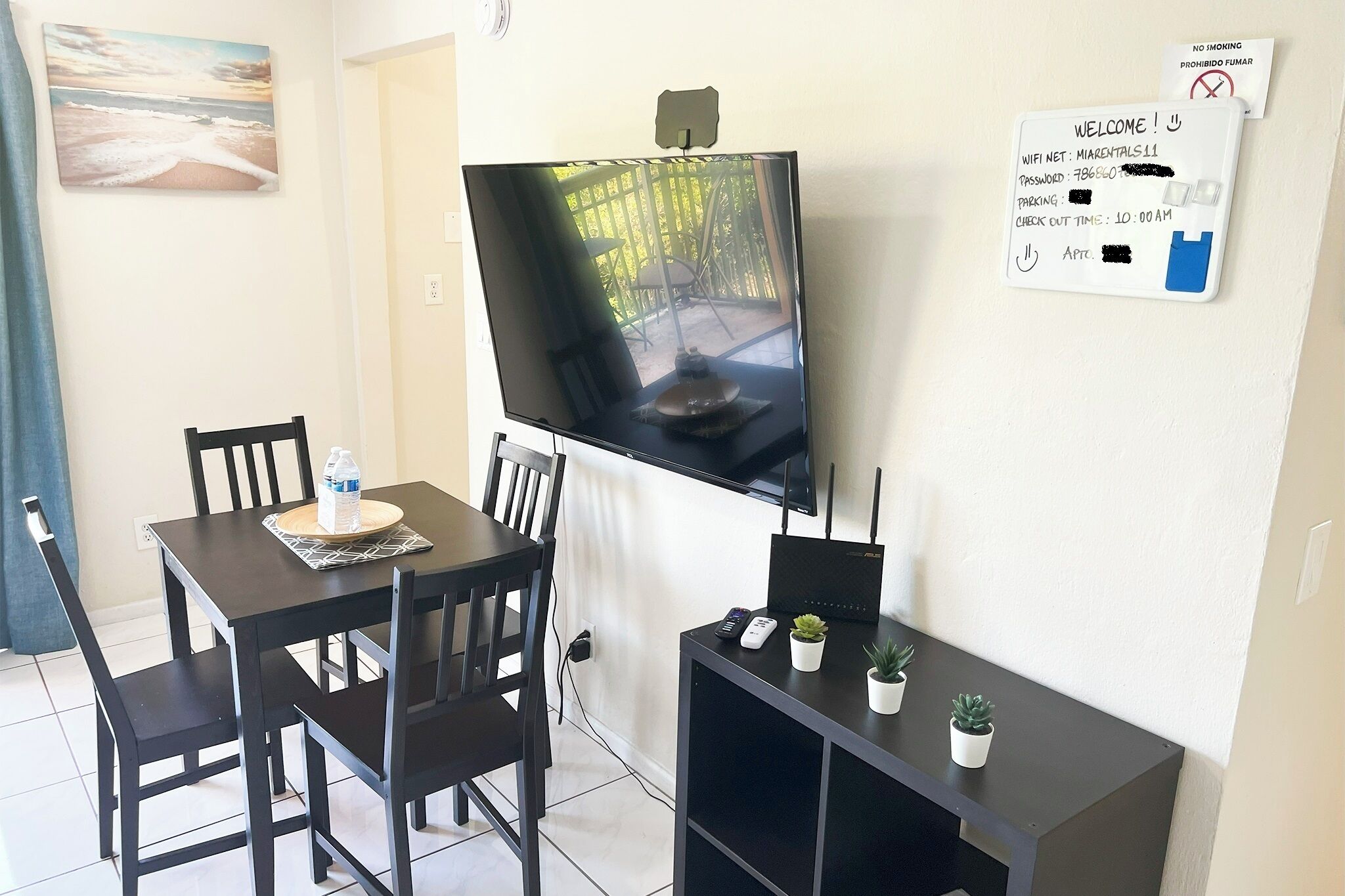 A room with a TV mounted on the wall, a table with chairs, a small plant on a side table, and a welcome sign with WiFi and parking information.
