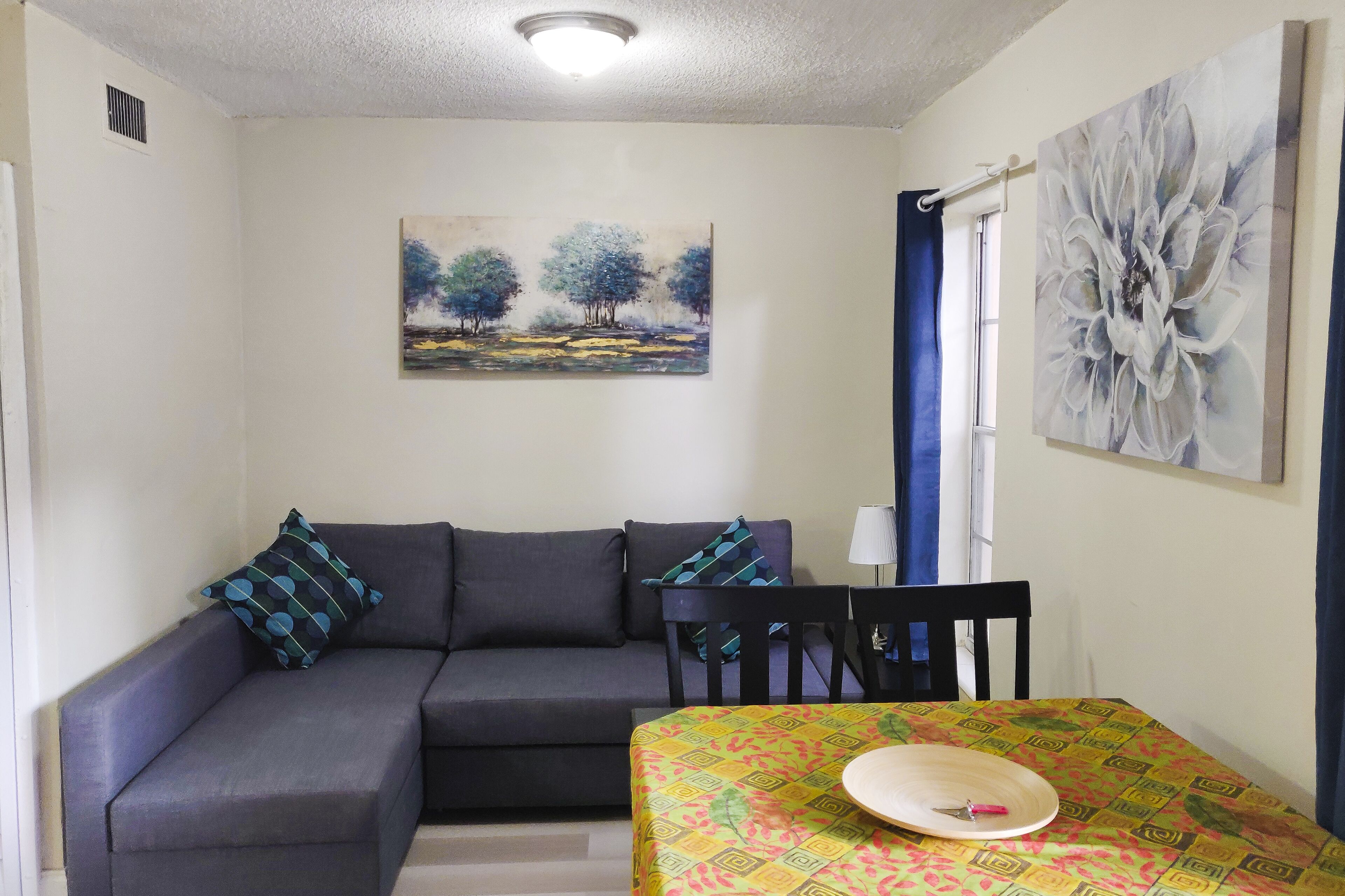 Una habitación con un sofá gris, una mesa de comedor con mantel estampado, un cuadro de árboles y un cuadro grande con motivos florales abstractos.