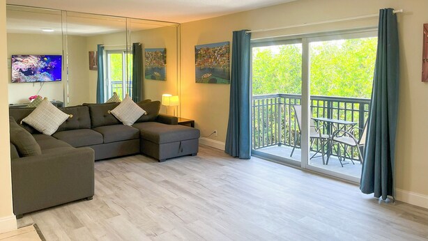 A modern living room with a sectional sofa, a flat-screen TV, a large mirror, and a balcony with outdoor furniture. - Sunny Isles Apartments by MiaRentals (Sunny Isles Beach)