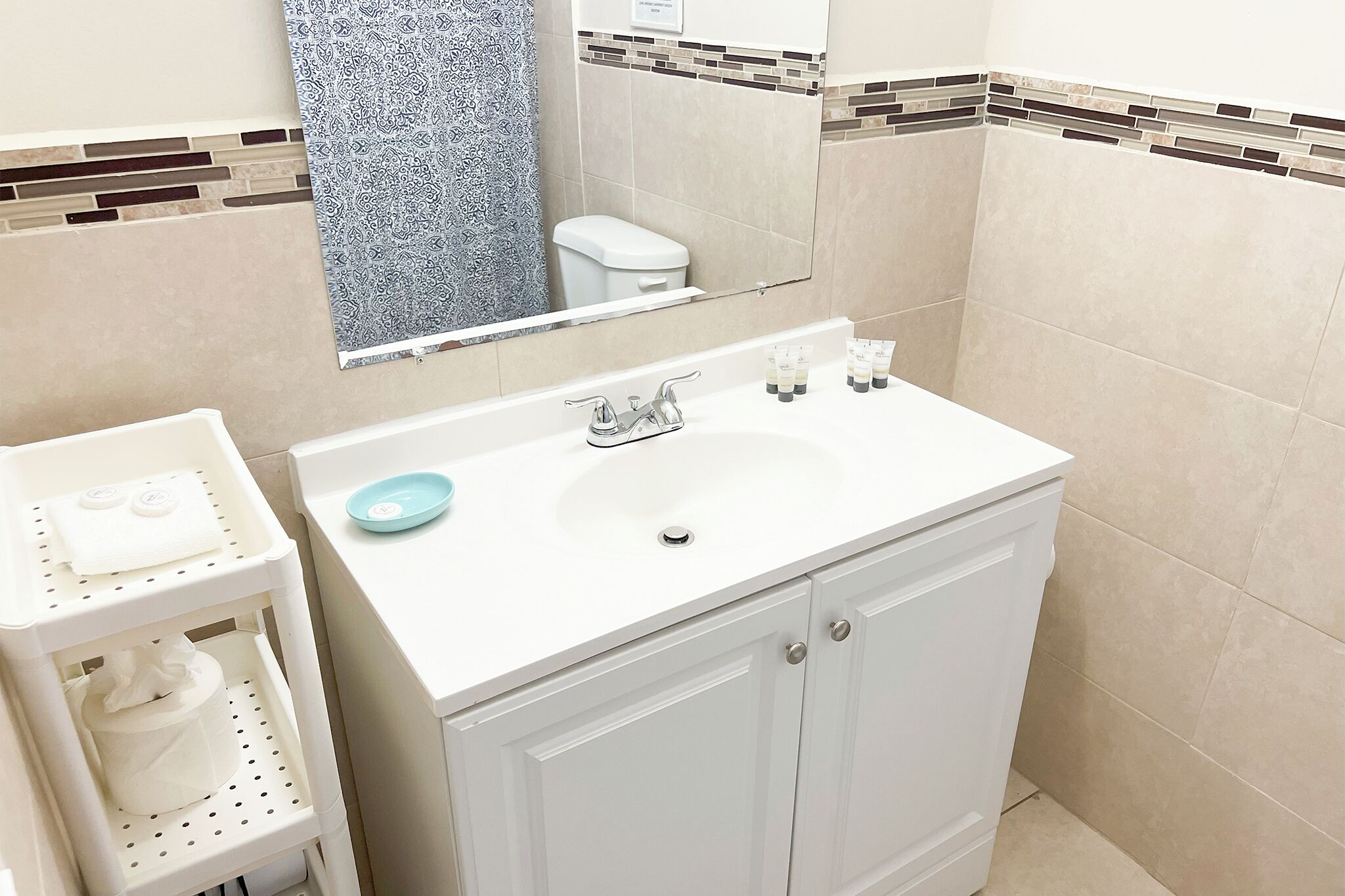 Un baño con lavamanos blanco, un espejo, inodoro y estante con artículos de aseo.