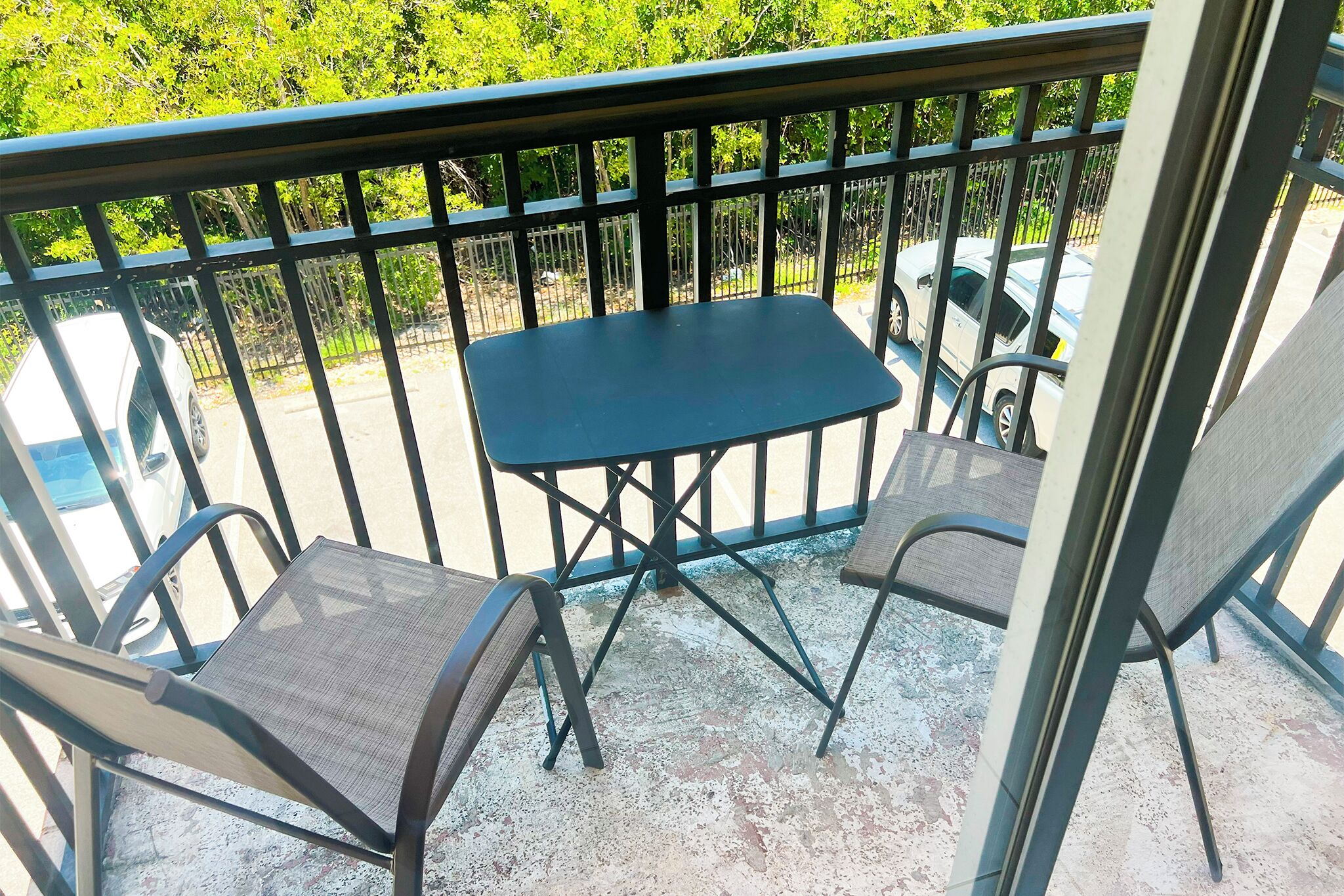A balcony with a table and two chairs, a metal railing, and a view of parked cars and greenery.
