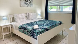 A neatly arranged bedroom with a bed, bedside tables, a lamp, a window with curtains, and a wall art piece. - Sunny Isles Apartments by MiaRentals (Sunny Isles Beach)
