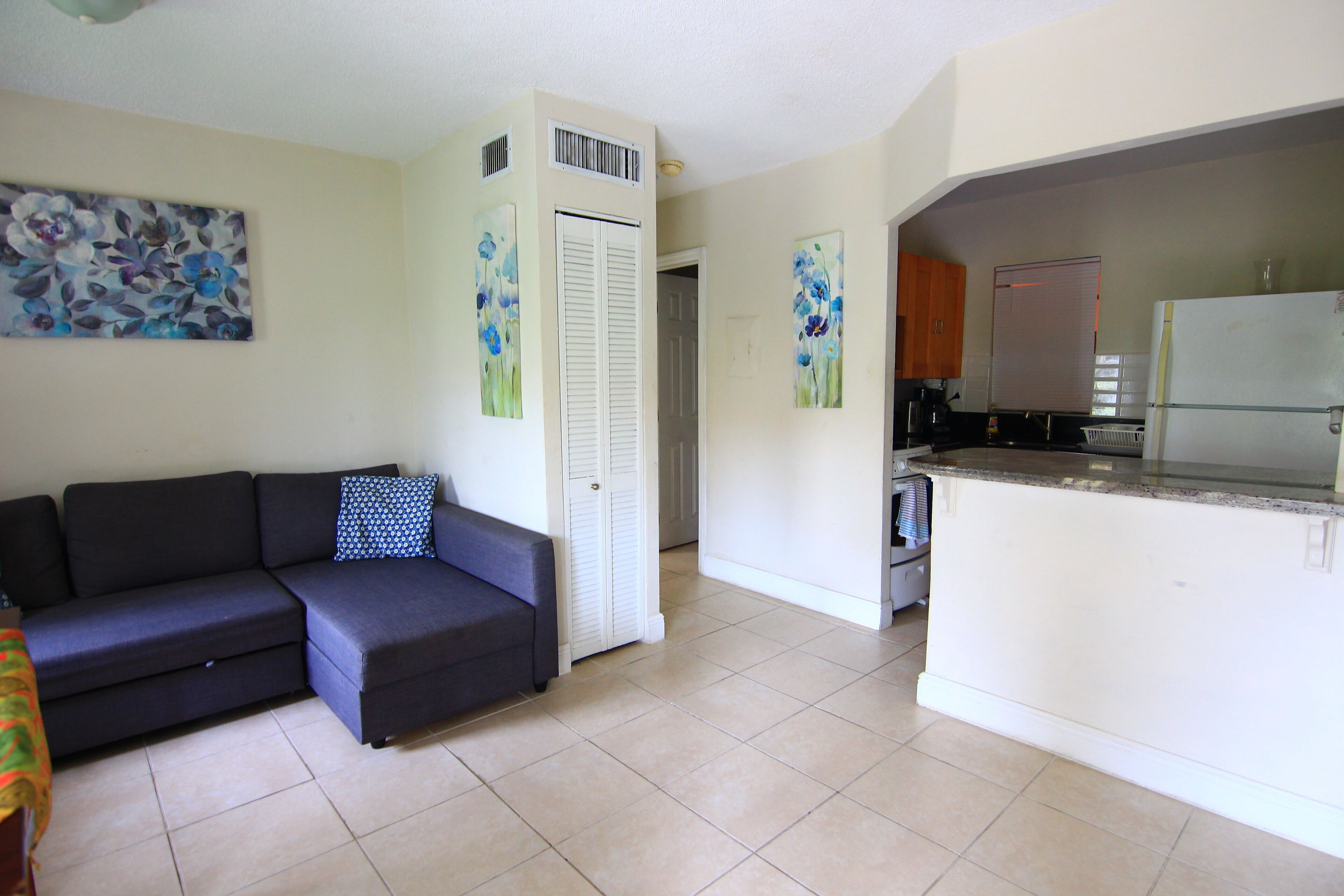 A living room with a sofa, a kitchen area with a refrigerator, and a door leading to another room.