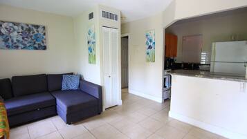 A living room with a sofa, a kitchen area with a refrigerator, and a door leading to another room.