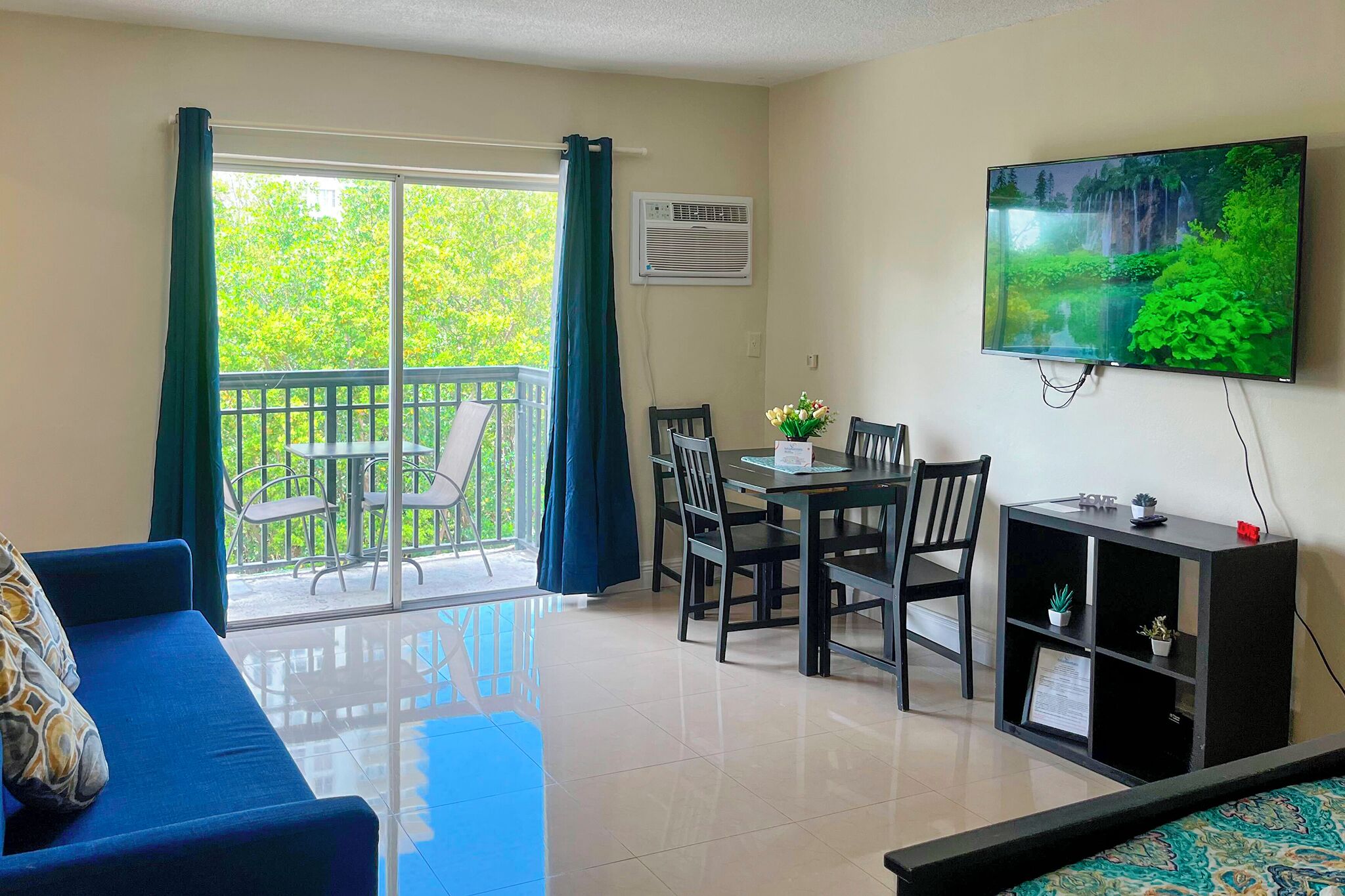 A living room with a blue sofa, a dining table with chairs, a TV on the wall, and a balcony with a view of trees.