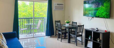 A living room with a blue sofa, a dining table with chairs, a TV on the wall, and a balcony with a view of trees.
