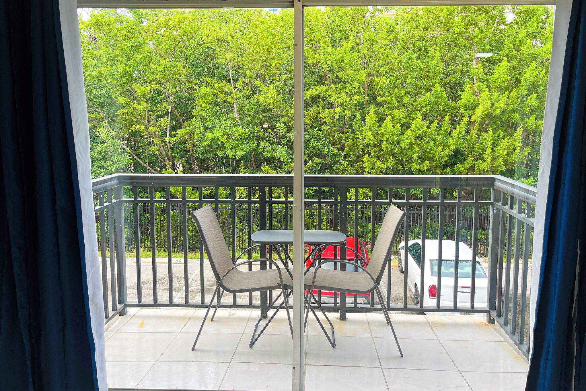 Un balcon avec deux chaises et une table, une balustrade, et une vue sur des arbres et une voiture garée.