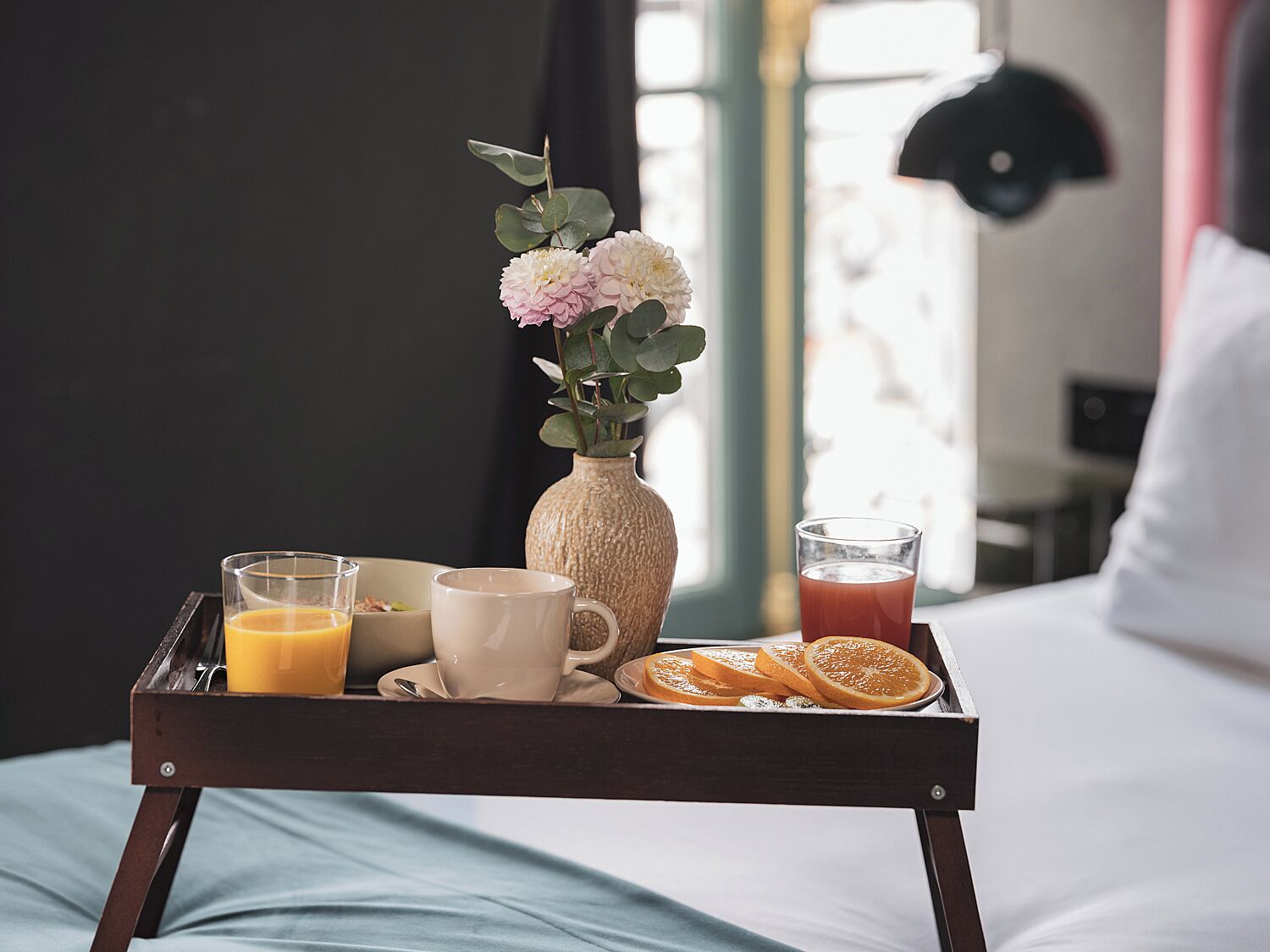 Una bandeja con jugo de naranja, tazas de café, rodajas de naranja y un florero con flores sobre una cama.