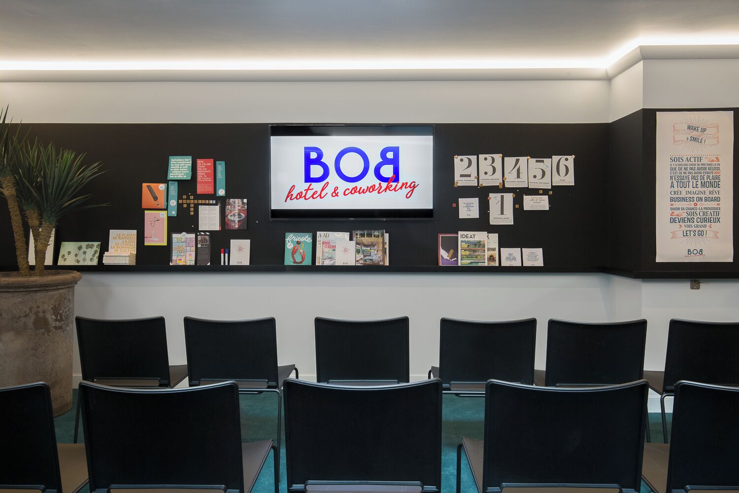 A hotel reception area with a large screen displaying the hotel's name, a bulletin board with various notices, and several black chairs arranged in rows.