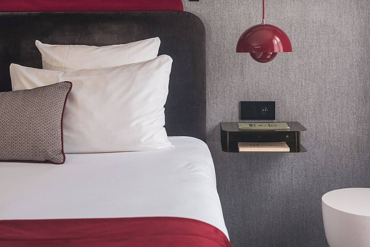 A modern hotel room with a large bed, a red accent wall, a red pendant light, and a small bedside table with a book.