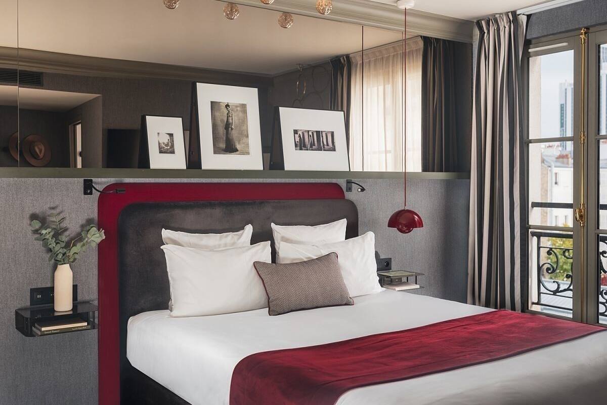 A hotel room with a bed, bedside tables, a mirror, artwork, and a view of the city.
