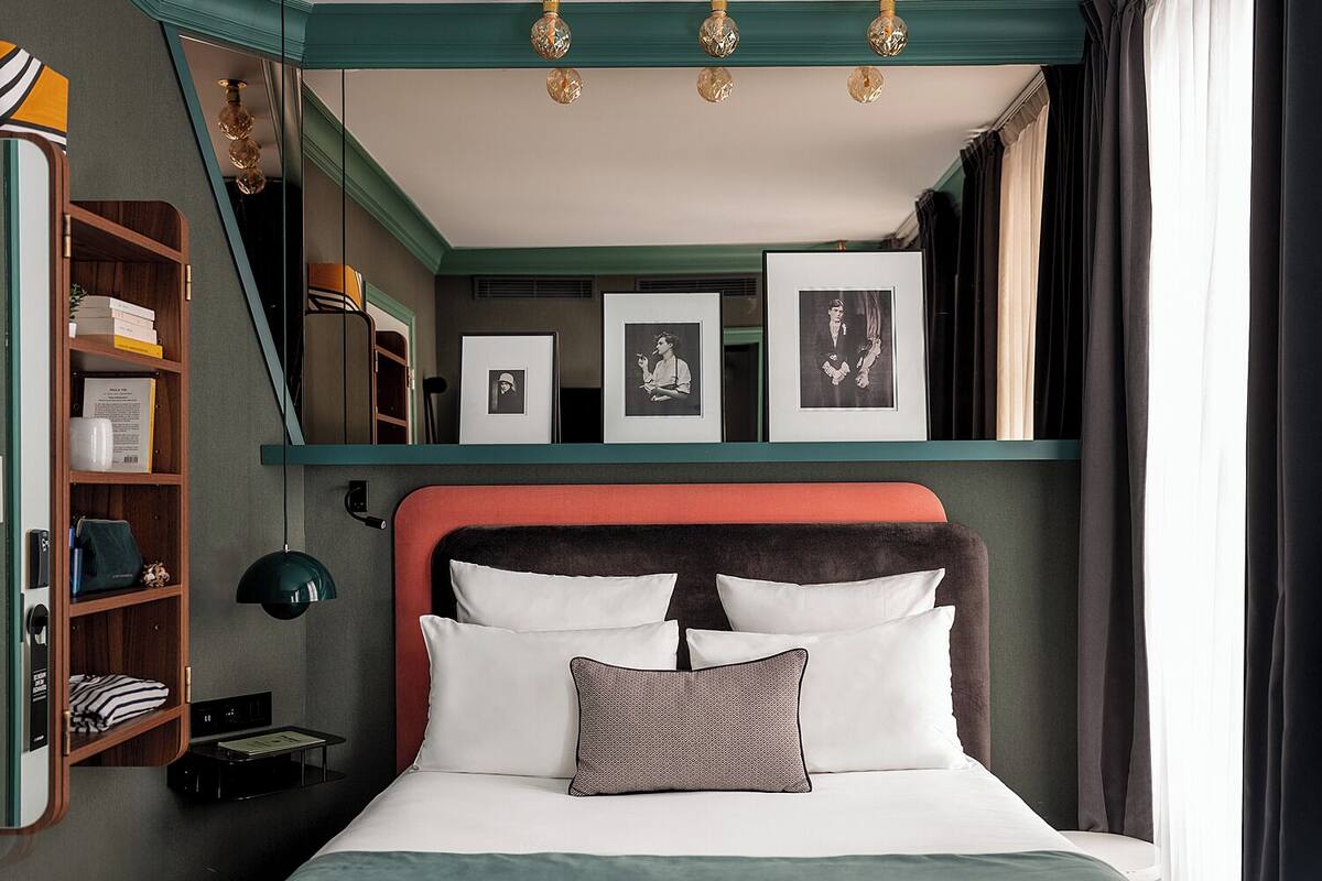 A neatly arranged bedroom with a bed, a shelf with books and framed photos, and a desk with a lamp.