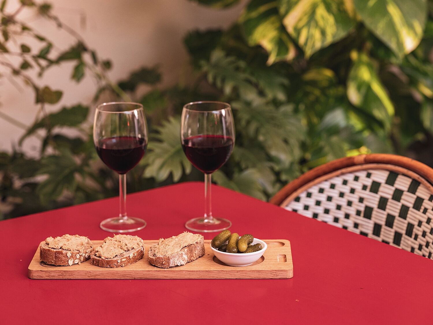 A table with two glasses of red wine, slices of bread, and a bowl of pickles on a red tablecloth.