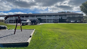 A motel with a two-story building, a parking lot with several cars, and a grassy lawn with a swing set. - Motel 6 Campbellford, ON (Trent Hills)