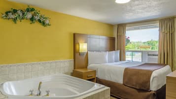A hotel room with a large bathtub, a bed with a brown blanket, a nightstand, a wall-mounted TV, and a window with a view of greenery.