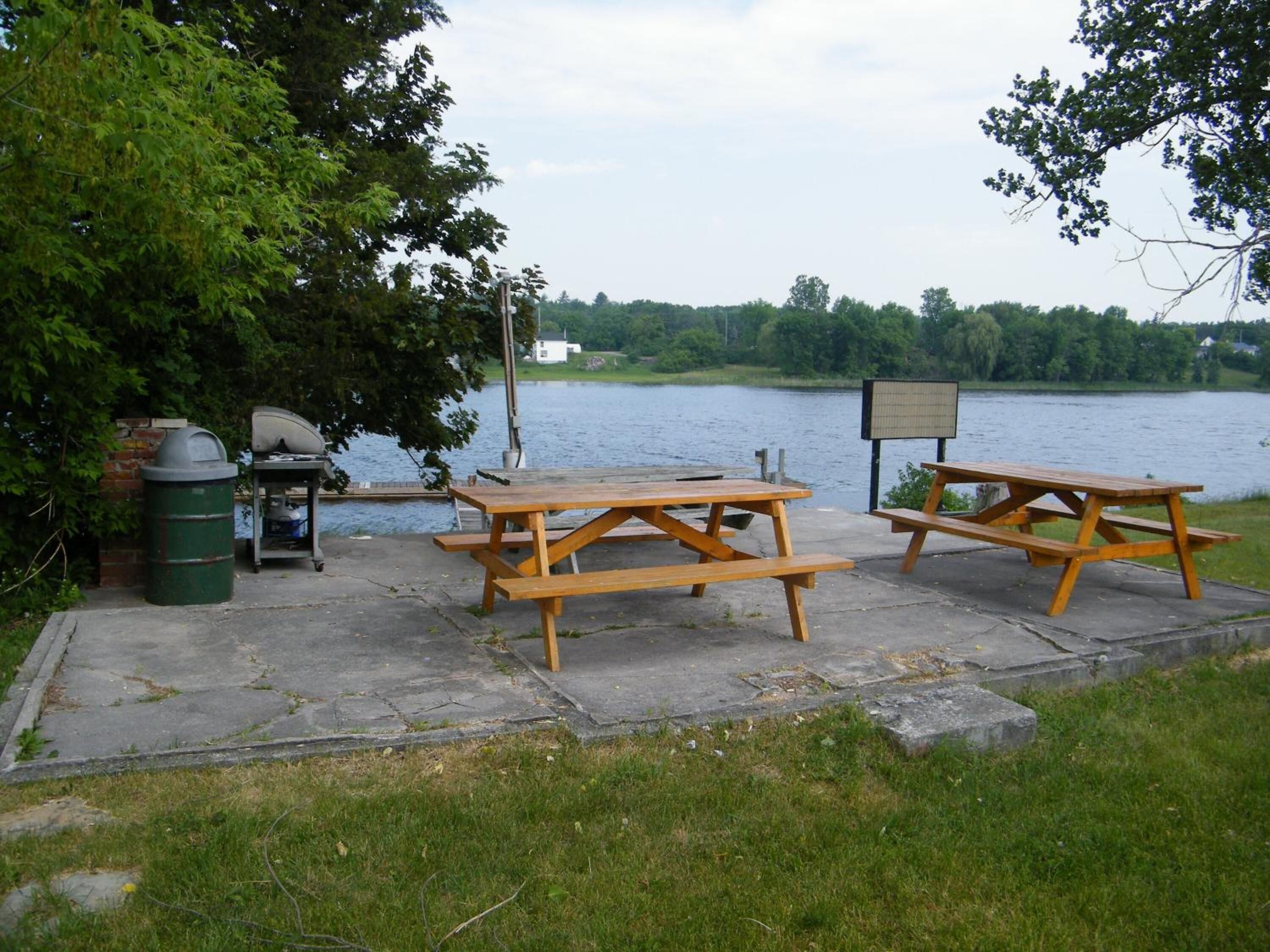 A picnic area with two wooden picnic tables and a trash bin, overlooking a lake.