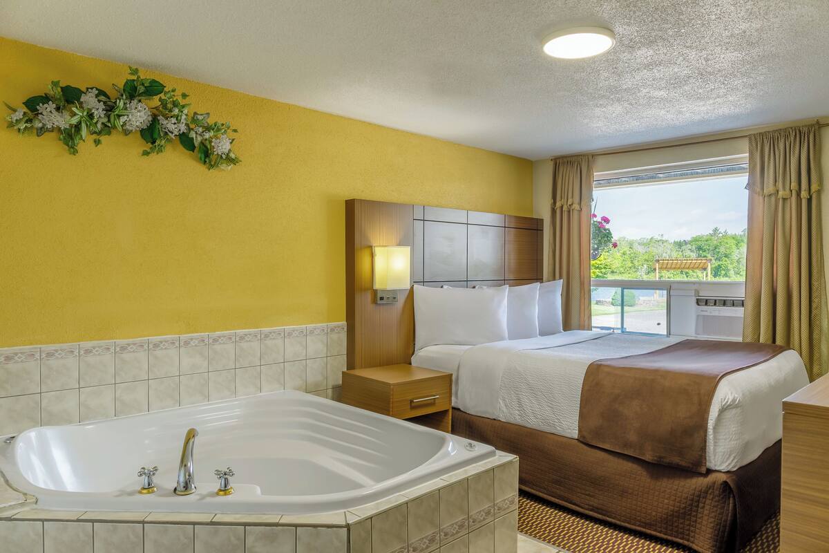 Habitación de hotel con una bañera grande, una cama con una manta marrón, una mesita de noche y vistas a zonas verdes.