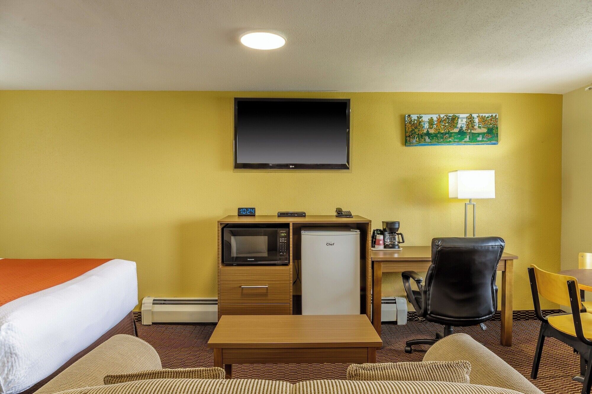 A hotel room with a bed, a desk with a computer, a refrigerator, a chair, a lamp, and a painting on the wall.