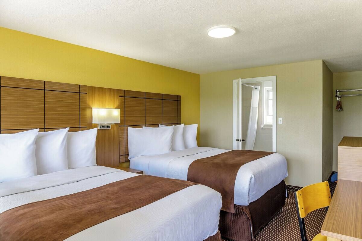Habitación de hotel con dos camas, cabecero de madera, mesita de noche con lámpara y baño visible a través de una puerta abierta.