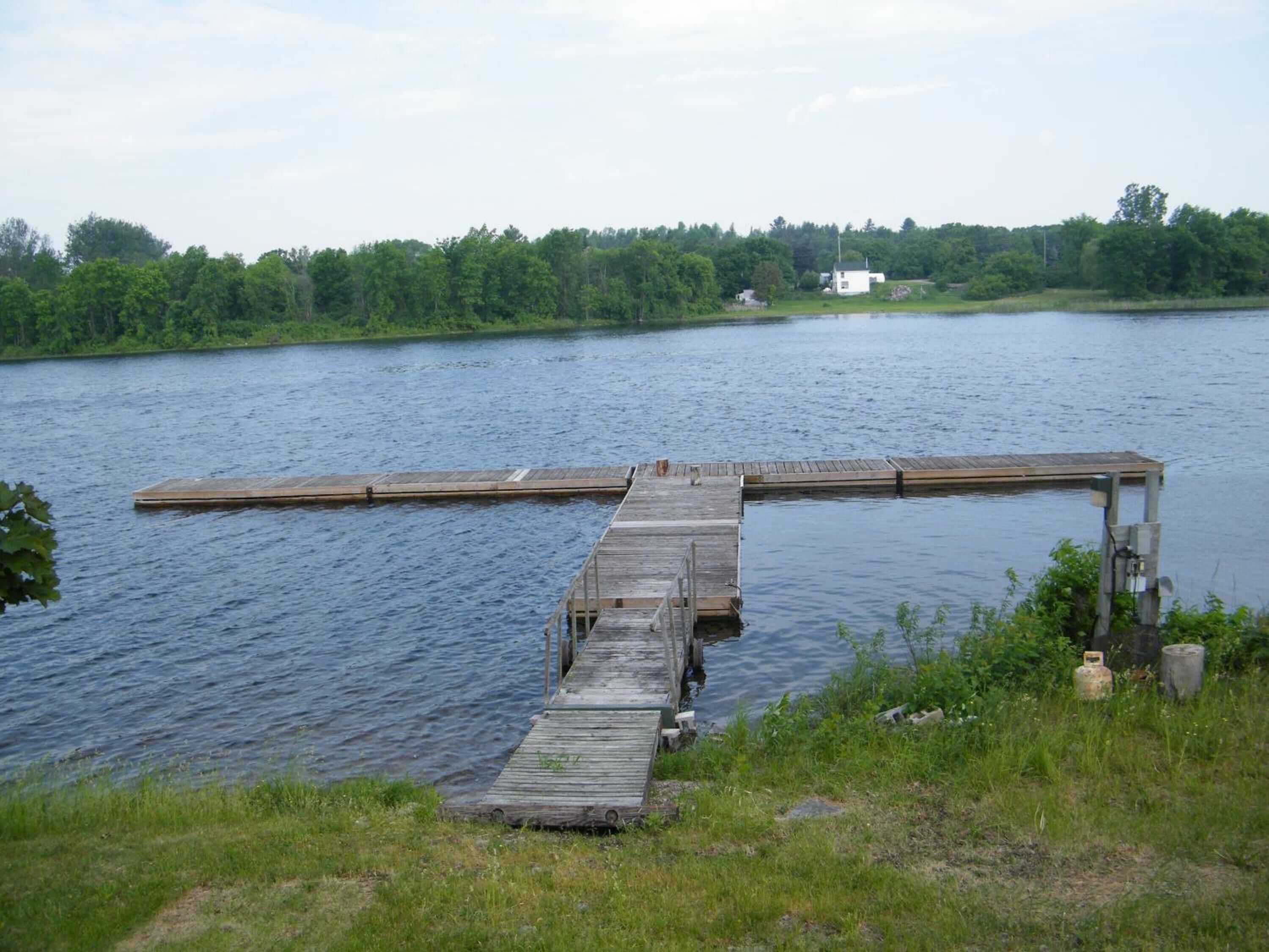 A wooden pier extending into a calm lake, surrounded by lush greenery and a distant white building.