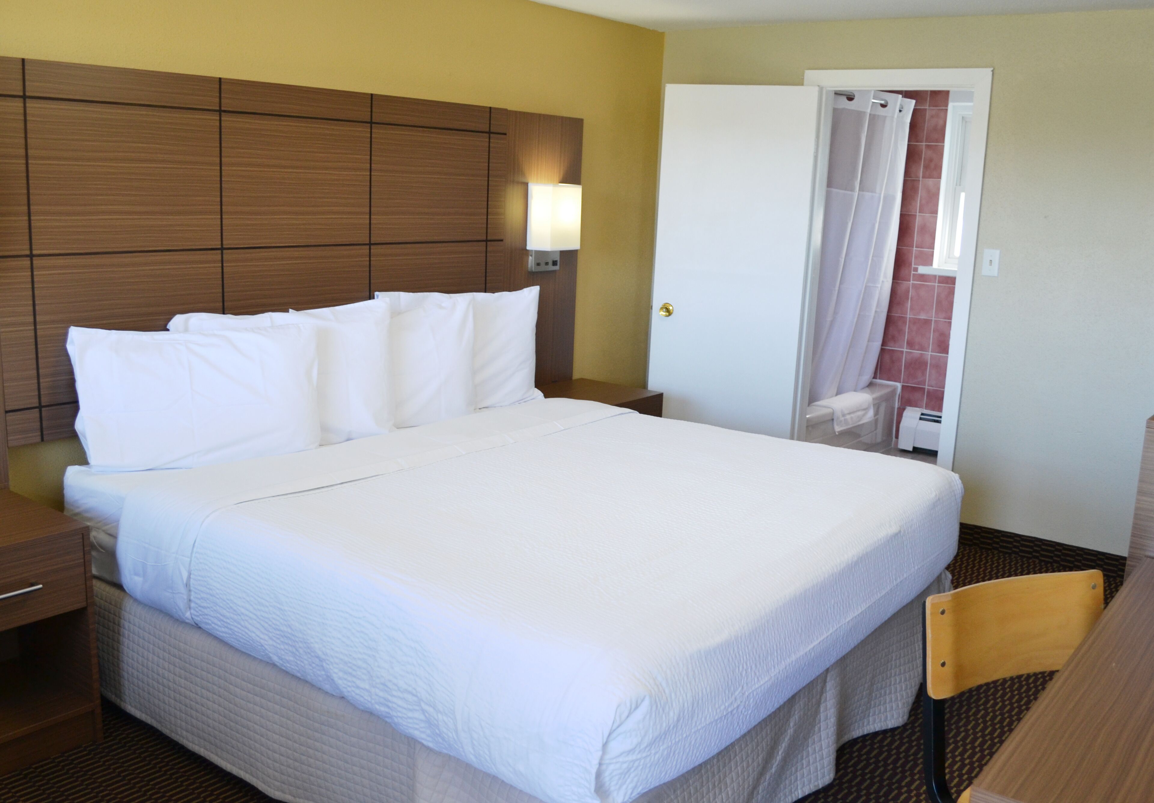 Habitación de hotel con una cama grande, cabecera de madera, mesita de noche, una silla y baño con cortina de ducha.