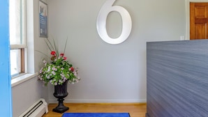 A room with a large white number '6' on the wall, a potted plant with flowers, and a blue rug on the floor. - Motel 6 Campbellford, ON (Trent Hills)