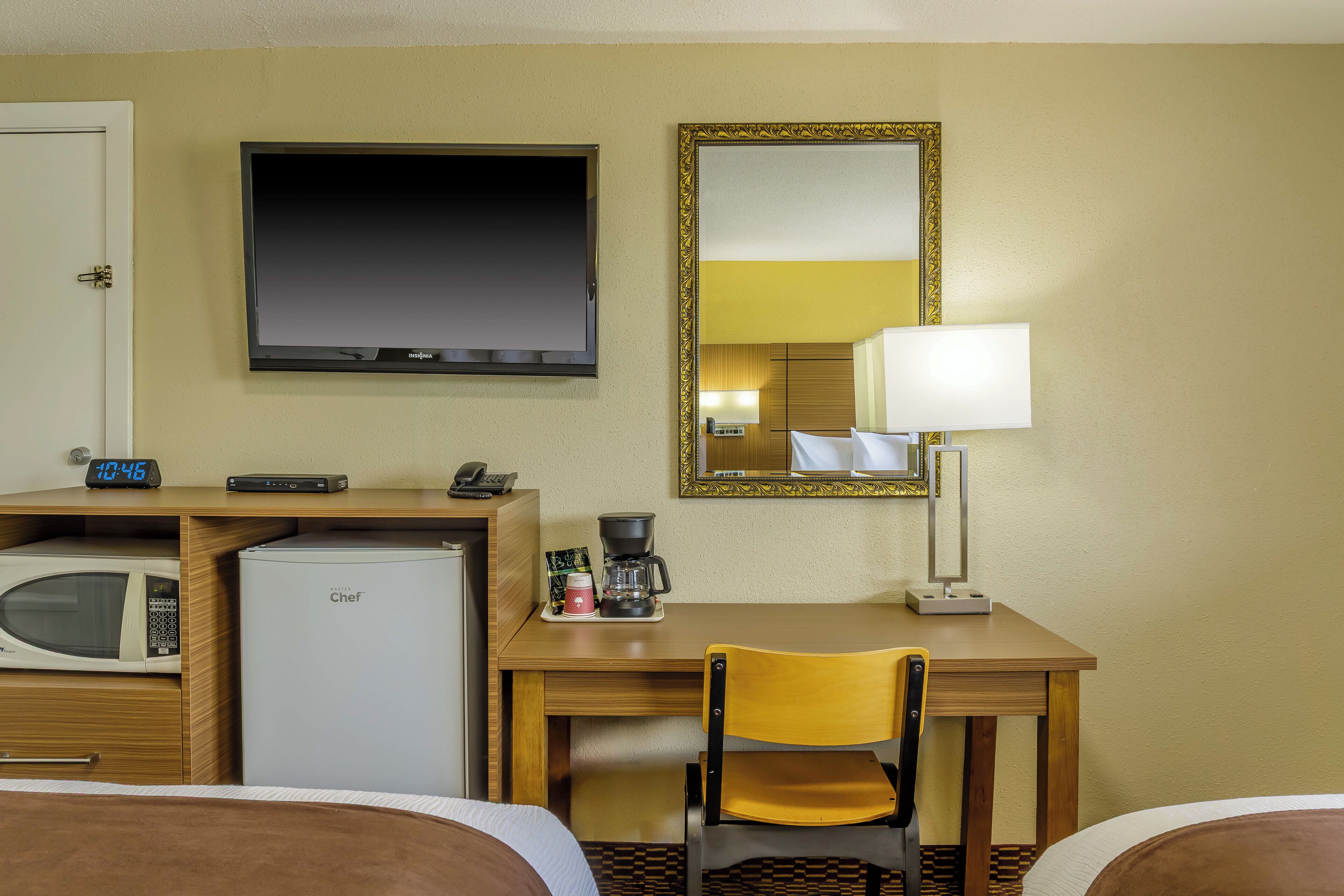 Ein Hotelzimmer mit einem Bett, einem Schreibtisch mit Stuhl, einer Mikrowelle, einem Kühlschrank, einem Fernseher, einer Lampe und einer Uhr.