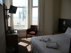 A hotel room with a bed, a chair, a desk with a monitor, and a window with curtains.