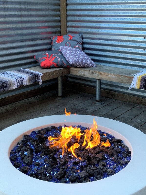A fire pit with a wooden bench and cushions in the background.