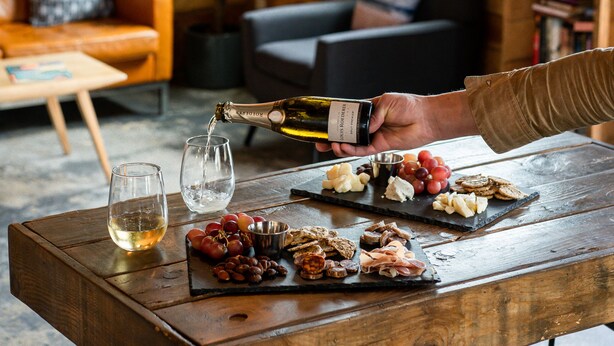 A wooden table with a wine bottle, glasses, and a charcuterie board in a cozy room with a wooden wall, a mirror, and a window. - Ashore Hotel (Seaside)