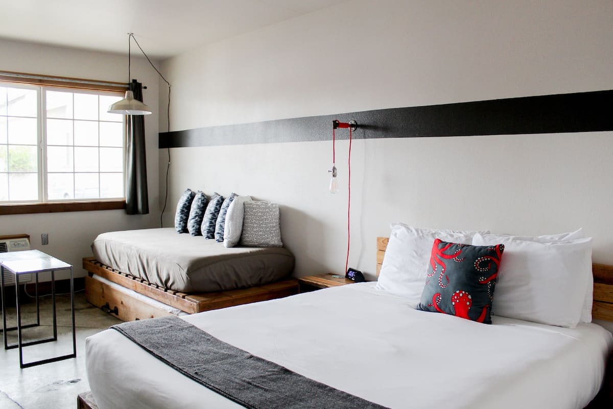 A double bed with a grey blanket, a bedside table, a lamp, and a window with blinds.