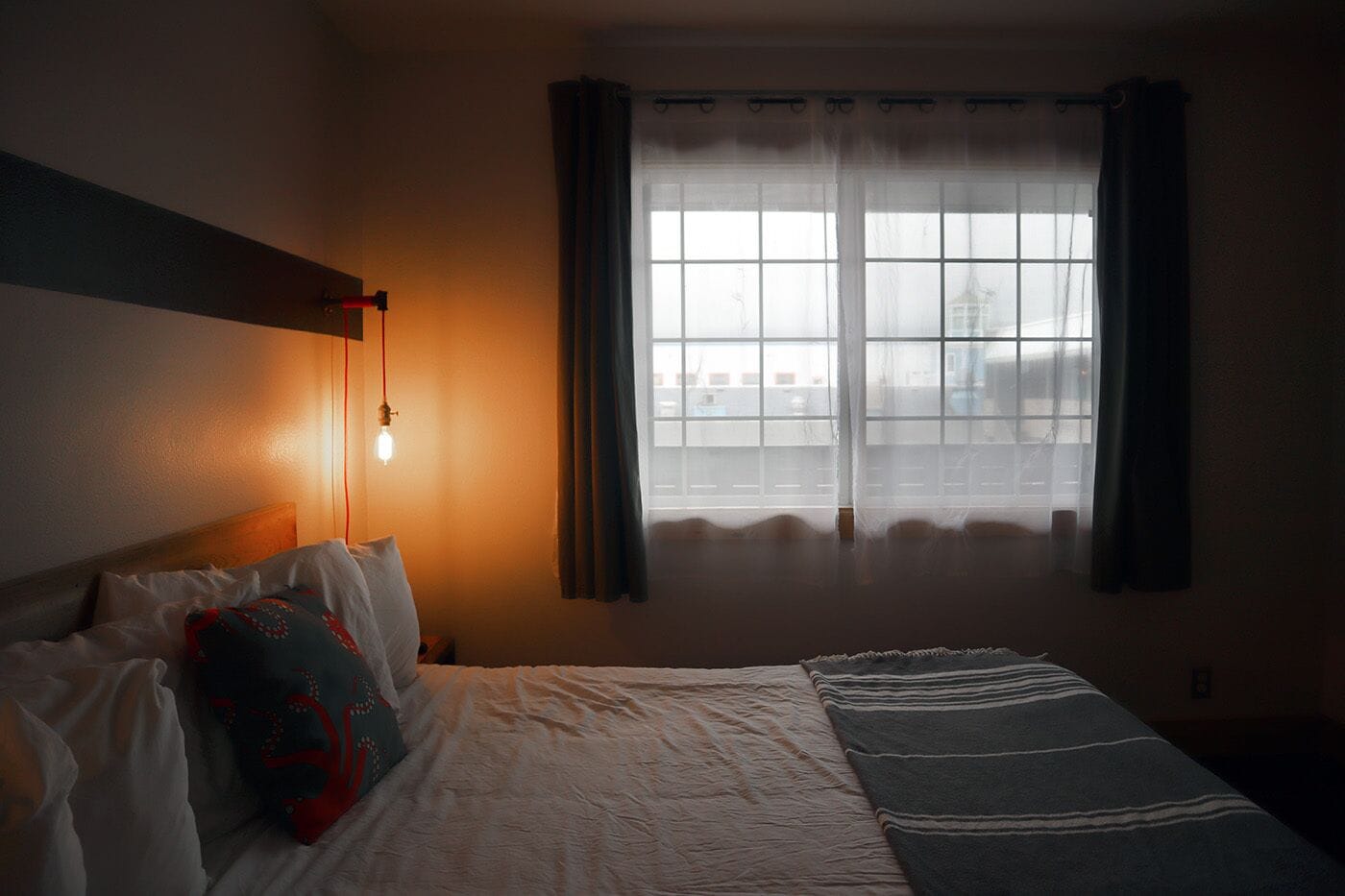 Ein Schlafzimmer mit Bett, Nachttischlampe, einem Fenster mit Vorhängen und Blick auf Gebäude.