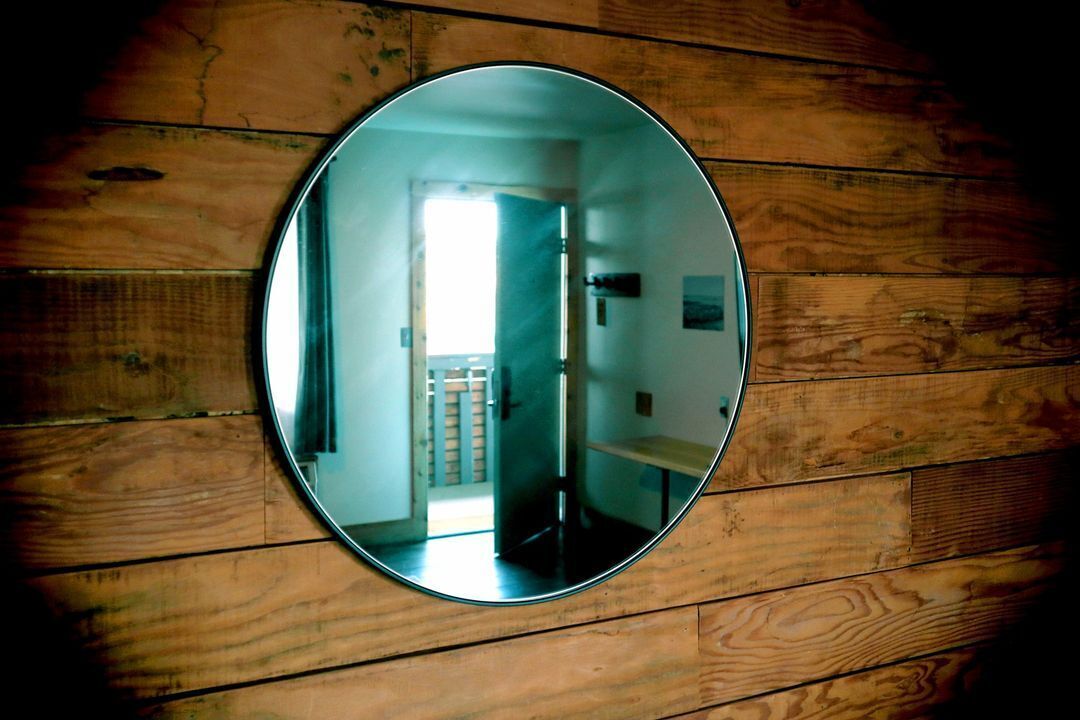 Un mirall rodó penjat a una paret de fusta que reflecteix una habitació amb una porta, un banc i una taula petita.