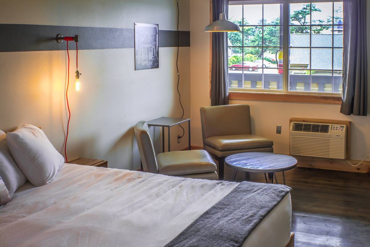A hotel room with a bed, a chair, a small table, a window with curtains, and an air conditioning unit.