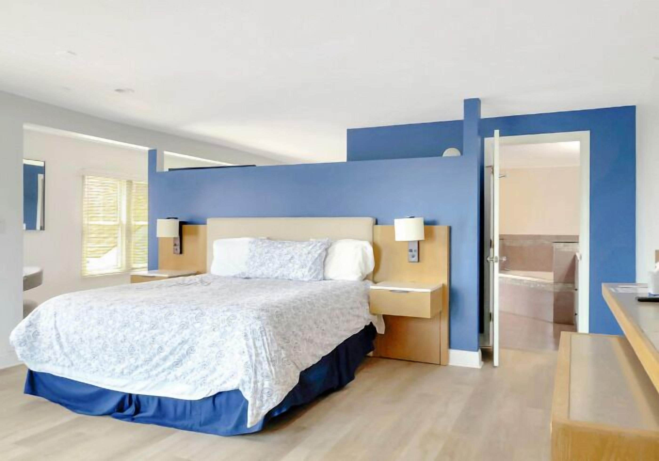 A modern hotel room with a large bed, bedside tables, a desk, and a bathroom visible through an open door.
