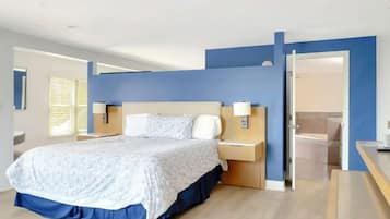 A modern hotel room with a large bed, bedside tables, a desk, and a bathroom visible through an open door.