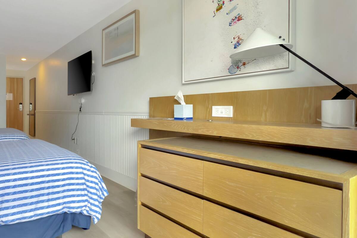 Habitación de hotel con cama, escritorio, silla, televisor montado en la pared y un cuadro enmarcado.