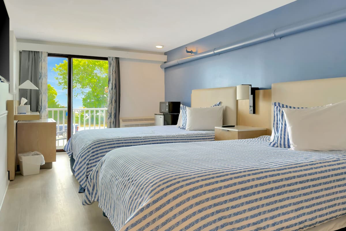 Habitación de hotel con dos camas, televisor, escritorio y balcón con vistas a árboles.